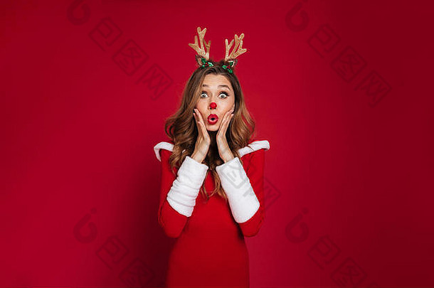 一个穿着圣诞鹿服装的惊艳美丽女孩的画像，她双手举在脸上，看着隔离在红色背景上的摄像机