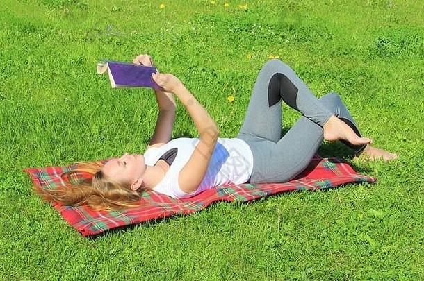 一个穿着白色T恤、留着长发的年轻漂亮的白人女孩躺在一块红色格子布上，躺在绿草上，躺在草坪上看书。