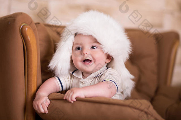 快乐的新生婴儿面颊粉红色的在黑格复古休闲风格的棕色沙发沙发沙发上摆姿势，沙发上戴着白色毛皮<strong>冬季保暖</strong>帽。