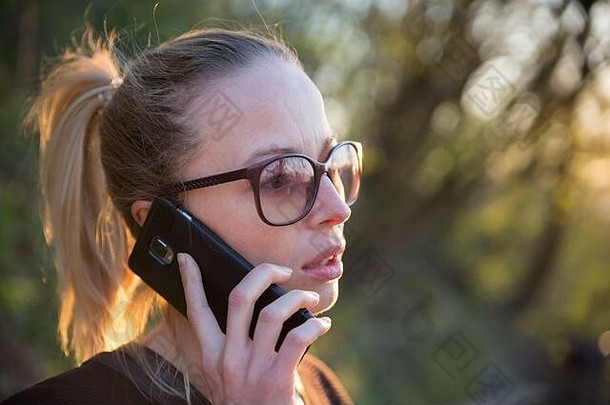 日落时分，一位年轻女子在公园户外打<strong>手机</strong>的背光后视图。女孩手持<strong>手机</strong>，使用<strong>数码</strong>设备，看着场景