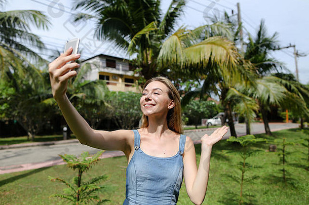 年轻漂亮的女孩在棕榈树旁用智能手机打视频电话。
