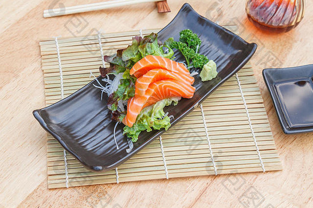 黑盘子里的刺身三文鱼配蔬菜和萝卜，木桌上放着筷子。