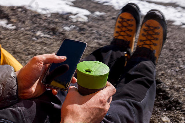一个登山者在雪山上用手机，手里拿着一个杯子。