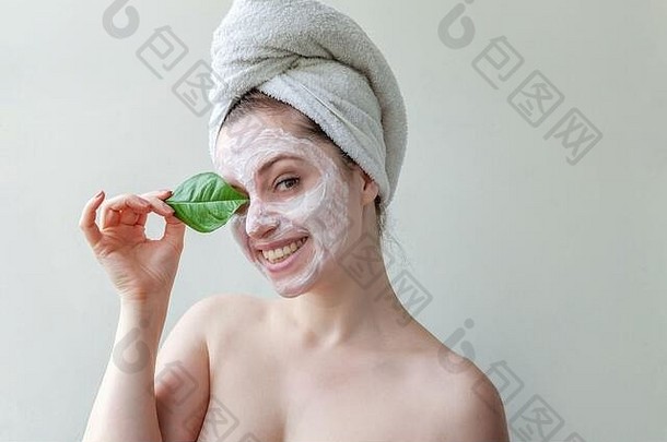 最低限度的美女肖像：头戴毛巾，脸上涂白色滋养面膜或润肤霜，手拿绿叶隔离白色背景。护肤清洁生态有机美容spa概念