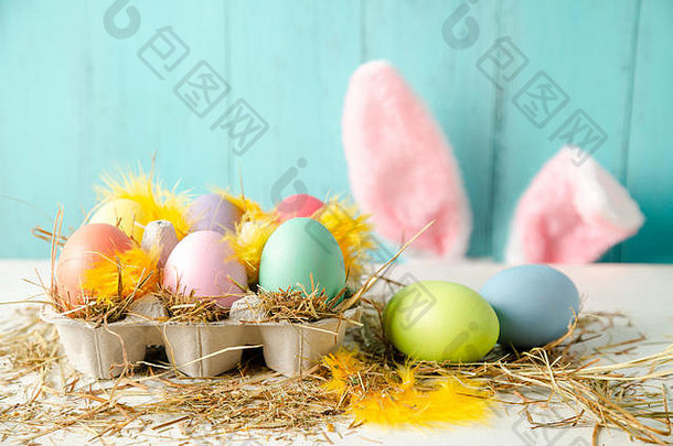 彩蛋盒中的彩色复活节彩蛋，以干草和羽毛为巢，后面是兔子耳朵，背景为木制绿松石色