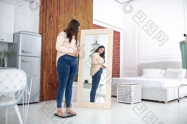 年轻的女人米色上衣站秤镜子