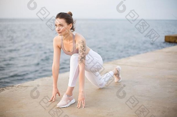 穿着<strong>运动</strong>上衣和白色紧身裤的沉思女孩练习瑜伽，背景为海景。一位年轻女子在河边训练时，梦幻般地将目光转向一边