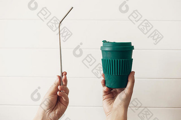 手持时尚的可重复使用的环保咖啡杯和白色木质背景上的钢吸管。零浪费。绿色杯由天然竹纤维和金属制成