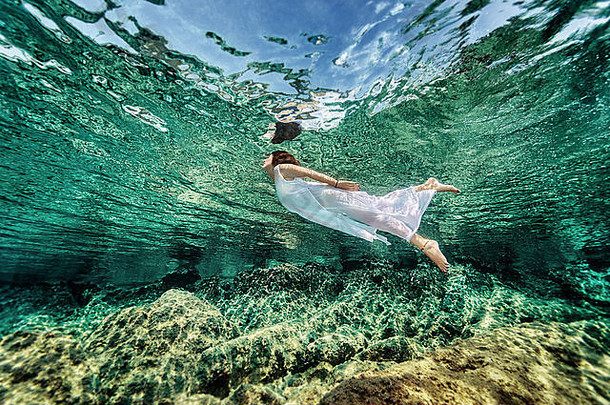 在透明的海洋中游泳，从清澈的海洋中浮出水面，穿着时尚的白色连衣裙，享受奢华的暑假
