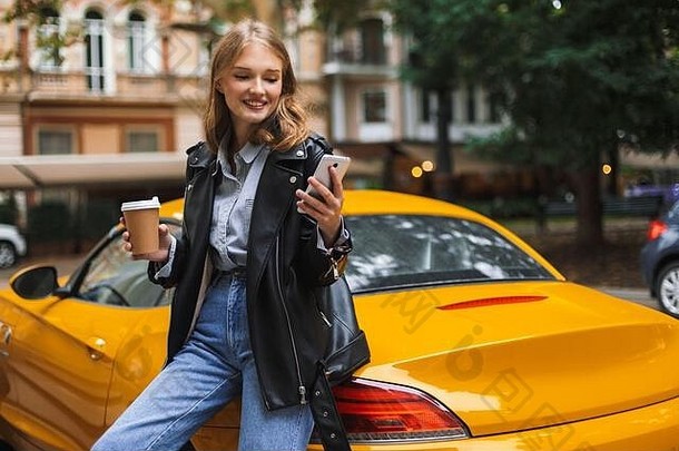 年轻的漂亮的微笑女人皮革夹克倾斜黄色的体育运动车持有杯咖啡手幸福的手机城市