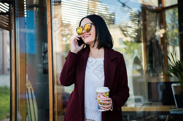 年轻的黑发女郎在城里闲逛，喝着咖啡。穿着外套，鲜红的嘴唇，对着咖啡馆的窗户摆姿势。模特一边看一边用手机聊天一边喝咖啡