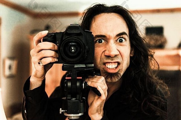 一位白人摄影师站在专业的单反相机和三脚架后面，脸上表情滑稽、令人毛骨悚然。