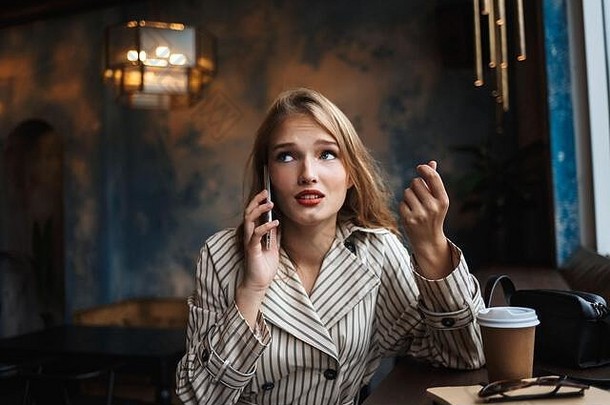 穿着条纹风衣的年轻女子一边在现代咖啡馆里消磨时光，一边用手机交谈，一边若有所思地抬起头，端着咖啡走近