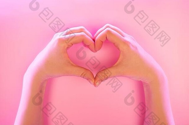 这位年轻女子的手在粉色背景上的时髦霓虹灯下做出了心形的手势。爱的概念。