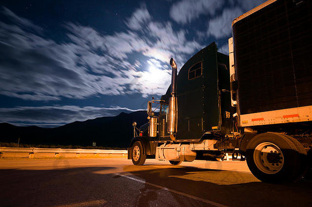 一辆黑色半<strong>卡车</strong>和一辆冷藏拖车在夜间停在停车场上的彩色照片，点亮了一盏灯笼，月光透过