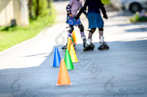 孩子们在路上用圆锥体学习溜冰。双胞胎女孩穿着gea防护服在家里的车道上练习安全轮滑
