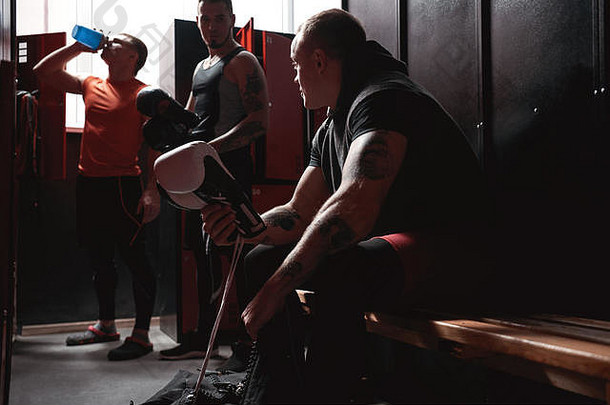 肌肉发达的运动员穿着运动服的拳击手为在黑暗的健身房更衣室训练准备彩色拳击手套