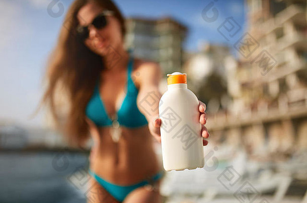 《快乐女人》在模糊的海滩背景上展示了防晒霜或乳白色的瓶子。皮肤黝黑的苗条女孩，穿着蓝色比基尼泳衣，手持