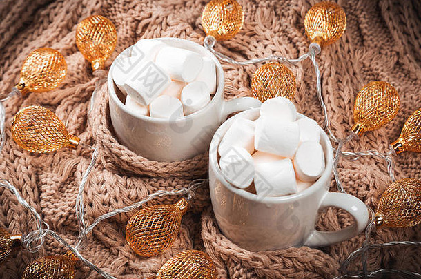 以可可或咖啡为背景，白色杯子里放着棉花糖，棕色针织冬季围巾，金色花环闪闪发光。漂亮的骗局