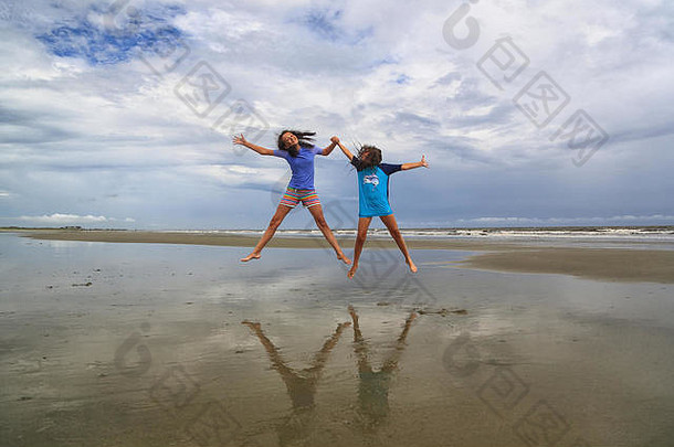 在南卡罗来纳州嘉华岛的海滩上，一个阴天，两个黑发女孩手拉手跳入空中。他们穿着游泳衣。
