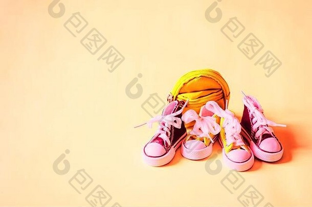 两双小运动鞋和黄色背包，在浅桃色背景上分开，空白处用于文本或设计。微型婴儿鞋和包。s