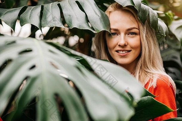 快乐迷人的白人金发女人，穿着红色t恤，在森林里享受时光，微笑着，牙齿洁白，站在热带树叶间。自然界