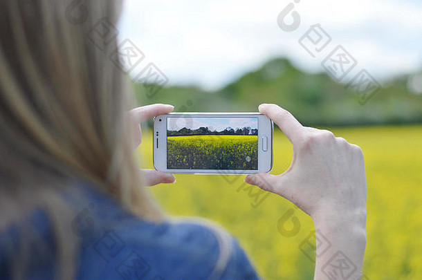 一个女人用智能手机拍了一张阳光灿烂的油菜田的照片。