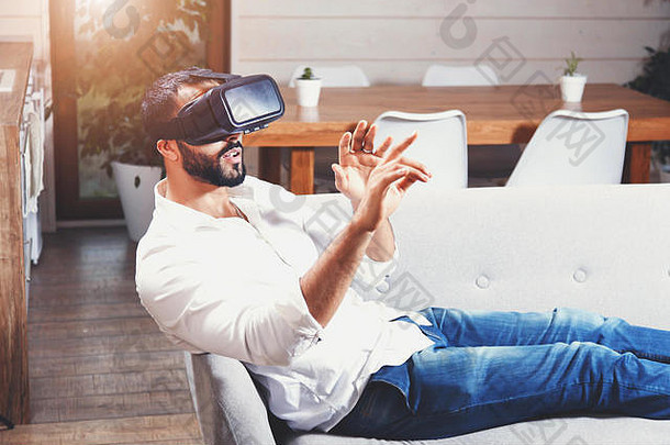 多元文化的胡子男人躺在沙发上，在现代公寓里使用虚拟现实护目镜