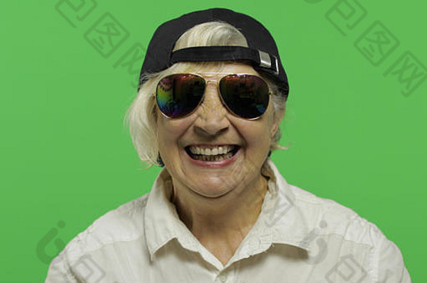 一位戴着墨镜和帽子的老妇人微笑着。穿着白衬衫的快乐老奶奶。放置您的徽标或文字。色度键。绿色屏幕背景