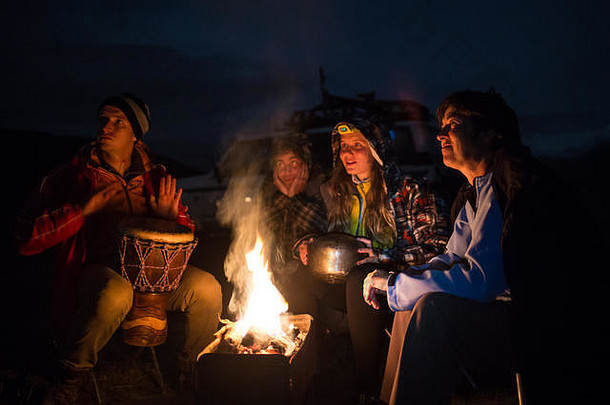 四个朋友坐在火炉旁，打鼓，在大自然中享受他们的露营生活。