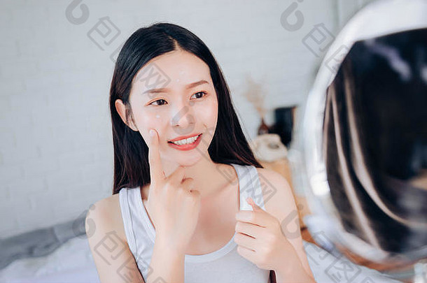 亚洲美女洁面后使用痤疮凝胶护肤品在白色卧室里看着镜子。