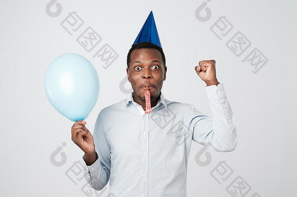 年轻的非洲男子穿着蓝色衬衫，戴着节日帽，吹着派对号角，在派对上玩得很开心。