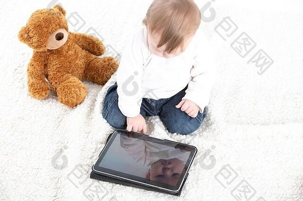 Stock studio照片，白色背景，一个婴儿看着平板电脑屏幕，旁边有一只泰迪熊。
