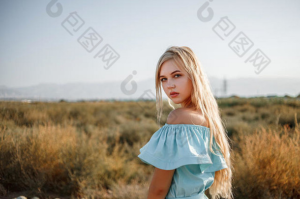 日落时，一位身穿浅蓝色连衣裙的年轻美丽的白人金发女孩站在<strong>西瓜地</strong>上，草<strong>地</strong>被晒干