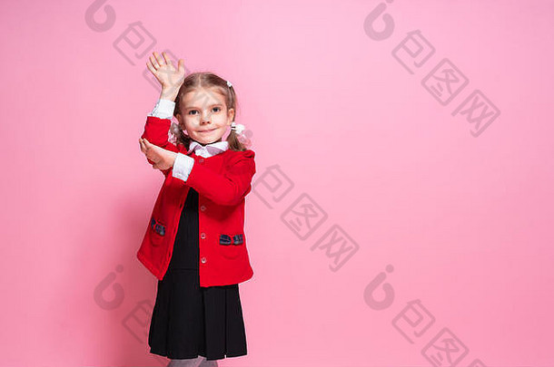 穿着鲜红色夹克和黑色校服的勤奋小女孩站在粉色背景上微笑着举起手