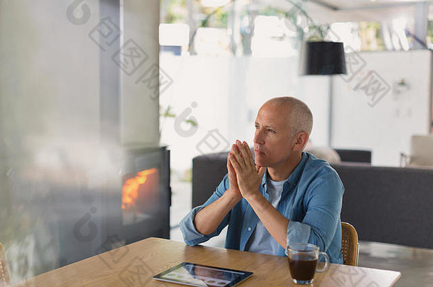 沉思的男人拿着数字平板电脑喝着咖啡，望着燃烧着木头的火炉壁炉