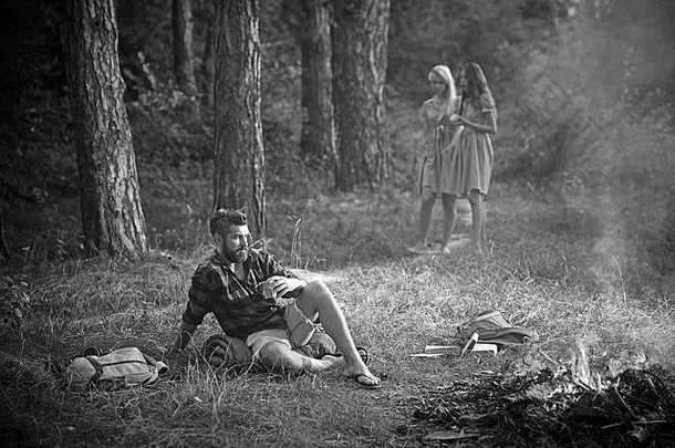 在森林里露营。男人一边喝茶一边看着火。那个家伙坐在阳光明媚的草地上。两个赤脚的女孩走在小路上。