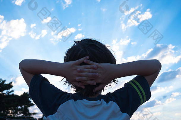 西班牙梅诺卡日落时仰望天空的11岁男孩的后视图，他伸出双臂，双手托着头
