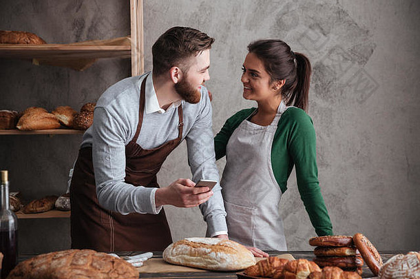 这对幸福的情侣面包师站在面包旁边，手里拿着手机。往旁边看。