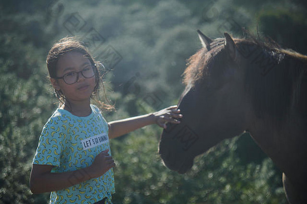 夏日阳光明媚，可爱的亚洲女孩站在野马环绕的田野里，抚摸着马头
