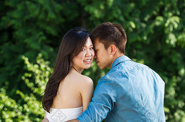 一对年轻的成年夫妇抱着对方，女朋友在她身后看着，背景是绿树