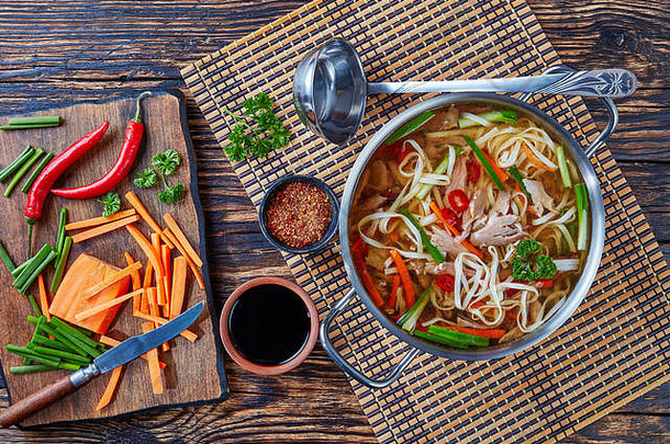 鸭面蔬菜汤放在一张古老的乡村餐桌上的金属砂锅里，上面放着勺子和食材，亚洲美食，俯瞰，flatlay