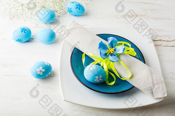 蓝色复活节餐桌，白色木质背景上有盘子、餐巾和蓝色装饰的复活节彩蛋