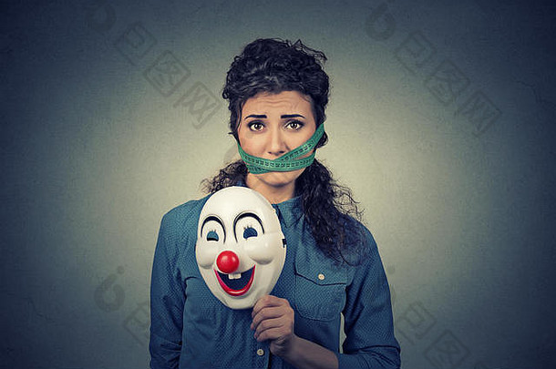 饮食限制和压力概念。在灰色墙壁背景上，戴着小丑面具、嘴上缠着卷尺的年轻悲伤女人的肖像。