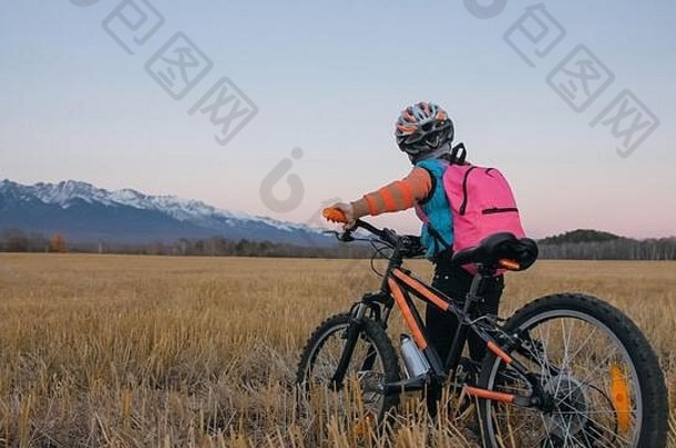 一个白人孩子在麦田里骑自行车。小女孩在美丽的雪山背景下走着<strong>黑橙</strong>色的自行车。自行车看台