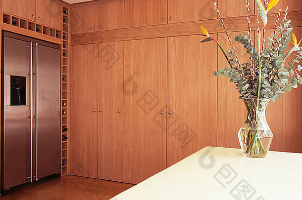 现代厨房的桌子上摆放着一瓶树叶和鲜花，配有大型木制<strong>储藏柜</strong>和美式冰箱