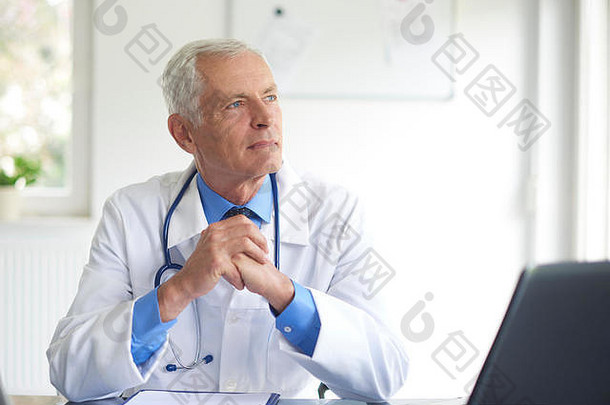 一张高级男医生坐在诊室的桌子旁沉思的画像。