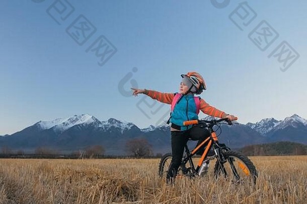 一个白人孩子在麦田里骑自行车。小女孩在美丽的雪山背景下走着<strong>黑橙</strong>色的自行车。