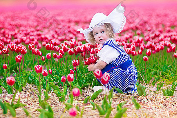 风车旁一片郁金香盛开的田野里，穿着荷兰传统民族服装的可爱卷发蹒跚学步的小女孩