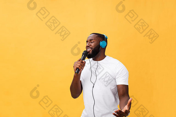 照片中是一位快乐、积极、时尚、英俊的非洲男子，他手持麦克风，头戴耳机，在黄色背景下听着<strong>音乐</strong>唱歌，享受周末假期